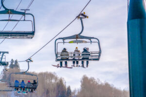 Skiers on ski lifts against sky in Park City Utah. Skiers enjoying a ride on chair lifts at a ski resort in Park City, Utah. Tall trees and bright cloudy blue sky can be seen in the background.